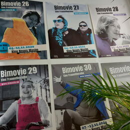 Sechs Plakate der Frauenfilmreihe Bimovie hängen in zwei Reihen an einer weißen Wand, teilweise von grünen Palmenblättern verdeckt. Jedes Plakat zeigt im oberen Bereich den Titel „Bimovie“ mit der jeweiligen Festivalnummer und der entsprechenden Homepage.